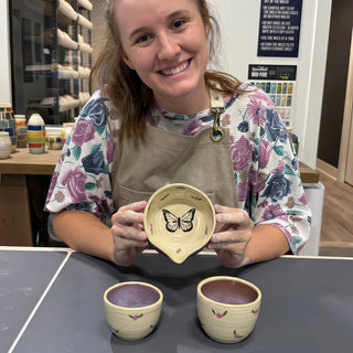 A member holding a small cream dish with a spout and black butterfly above two small flowered cups at The Clay Hole pottery studio in Draper, Utah