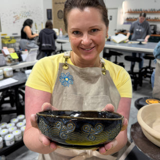 A member holding out a dark glossy bowl with carved swirls and wave lines around its sides at The Clay Hole pottery studio in Draper, Utah