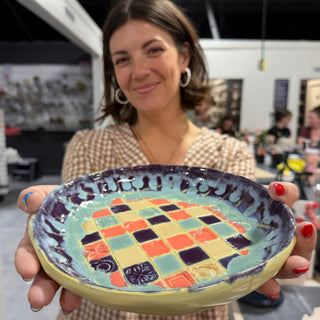 A member holding a shallow dish with a checkerboard of coral, teal, yellow and purple squares and a purple rim at The Clay Hole pottery studio in Draper, Utah
