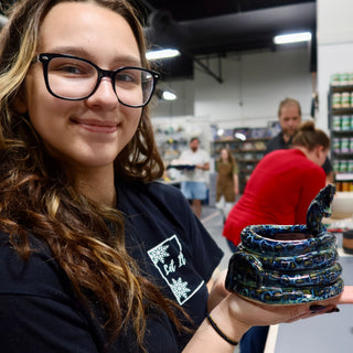 A member holding a coiled snake-shaped bowl with a raised head and dark blue and green glossy glaze at The Clay Hole pottery studio in Draper, Utah
