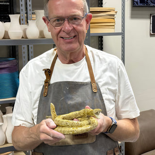 A member in a gray apron holding a coiled yellow-green snake sculpture with a raised head and open mouth at The Clay Hole pottery studio in Draper, Utah