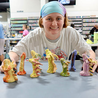 A member leaning over a table with several small standing creature figures in orange, yellow, green, blue and purple glazes at The Clay Hole pottery studio in Draper, Utah