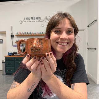 A member cupping a wide copper orange mug with a looped handle and a sculpted nose on the front at The Clay Hole pottery studio in Draper, Utah