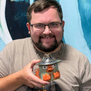 A member holding a small round figurine wearing a hat, with wide eyes, an orange beak and orange feet at The Clay Hole pottery studio in Draper, Utah