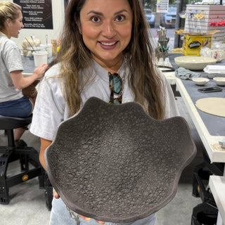 A member holding a wide dark gray dish with a wavy scalloped edge and a dotted texture at The Clay Hole pottery studio in Draper, Utah