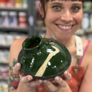 A member holding a squat dark green vessel with a pale curved line and a small black roof shape at The Clay Hole pottery studio in Draper, Utah