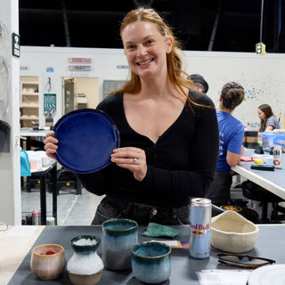 A member holding a round deep blue plate, with small glazed cups and a pale dish on the table at The Clay Hole pottery studio in Draper, Utah