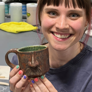 A member holding a rust brown mug with a green inside and a sculpted nose and mustache on the front at The Clay Hole pottery studio in Draper, Utah