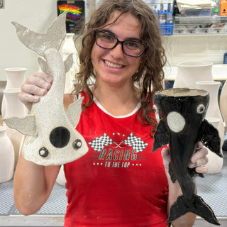 A member holding two fish shaped vessels, one speckled white with a black eye and one glossy black at The Clay Hole pottery studio in Draper, Utah