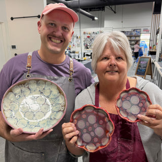 Two members holding a large plate with a pale flower-like pattern and two small scalloped pink dishes at The Clay Hole pottery studio in Draper, Utah