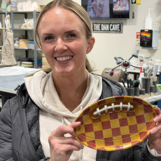 A member holding an oval platter shaped like a football with a red and yellow checkerboard pattern and white laces at The Clay Hole pottery studio in Draper, Utah