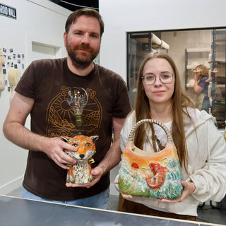 Two members holding a fox figure topped with a light bulb and a handbag-shaped piece with a fish in waves at The Clay Hole pottery studio in Draper, Utah