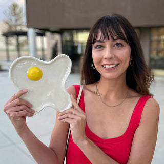 A member holding a white dish shaped like a fried egg with a bright yellow yolk in the center, with The Clay Hole, a pottery studio in Draper, Utah