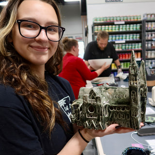 A member holding a gray-green sculpture of a building with two tall spires and arched windows at The Clay Hole pottery studio in Draper, Utah