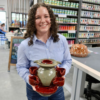 A member holding a gray-green vessel with a red glossy base and several small red cups around its sides at The Clay Hole pottery studio in Draper, Utah