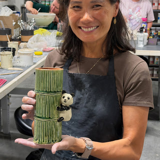 A member in a dark apron holding a tall green bamboo-shaped vessel with a small panda figure on its side at The Clay Hole pottery studio in Draper, Utah