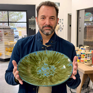 A member holding a large shallow bowl with radiating green streaks, a blue speckled center and a blue rim at The Clay Hole pottery studio in Draper, Utah