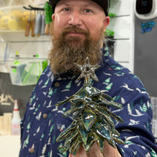 A member holding a small glossy green tree figurine with layered branches and a star on top at The Clay Hole pottery studio in Draper, Utah