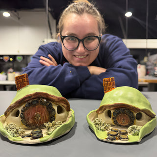 A member leaning on folded arms behind two green domed cottage sculptures with round wooden doors and brick chimneys at The Clay Hole pottery studio in Draper, Utah