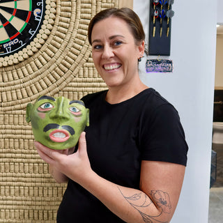 A member holding a green ceramic face with painted eyes, a black mustache and an open mouth showing teeth at The Clay Hole pottery studio in Draper, Utah