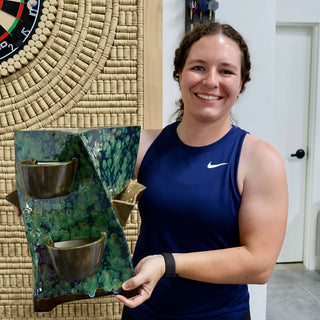 A member holding a tall folded green panel with mottled glaze and two brown bowl-shaped cups attached at The Clay Hole pottery studio in Draper, Utah