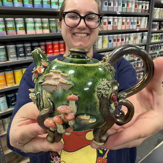 A member holding a glossy green teapot covered in clay mushrooms, with a curled brown handle and curled brown feet at The Clay Hole pottery studio in Draper, Utah