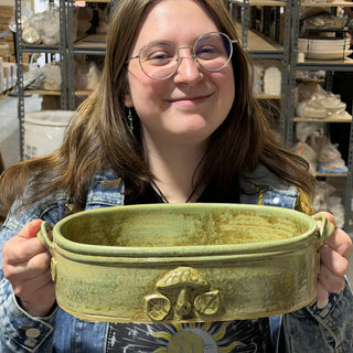 A member holding a long oval green dish with two side handles and a raised mushroom decoration on the front at The Clay Hole pottery studio in Draper, Utah