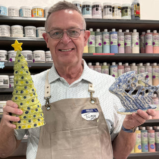 A member holding a tall yellow green cone tree with blue dots beside a blue and white sculpture with holes at The Clay Hole pottery studio in Draper, Utah