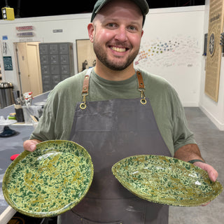 A member holding two oval plates covered in dark green and olive speckles at The Clay Hole pottery studio in Draper, Utah
