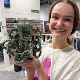 A member holding a tall green and black speckled vase with interlocking ring shapes along its sides at The Clay Hole pottery studio in Draper, Utah