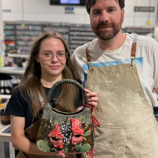 Two members standing together, one holding a handbag-shaped piece with a braided handle, red horned masks and green swirls at The Clay Hole pottery studio in Draper, Utah