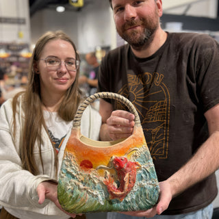Two members holding a handbag-shaped piece with a braided handle, a raised red fish in waves and an orange sky at The Clay Hole pottery studio in Draper, Utah