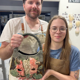 Two members showing a handbag-shaped piece with two red horned faces, a pale flower between them and a braided handle at The Clay Hole pottery studio in Draper, Utah