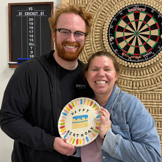 Two members holding a white plate that says Happy Birthday with a blue cake and colorful dashes around the rim at The Clay Hole pottery studio in Draper, Utah
