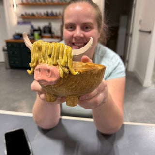 A member smiling behind an amber bowl shaped like a shaggy cow with a pink snout and cream horns at The Clay Hole pottery studio in Draper, Utah