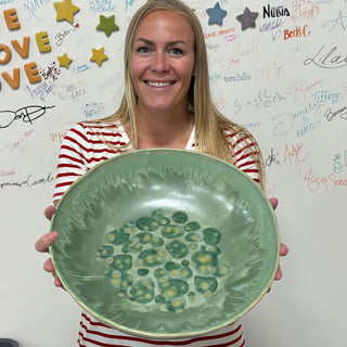 A member holding a large shallow bowl in soft green glaze with darker green spots in the center at The Clay Hole pottery studio in Draper, Utah