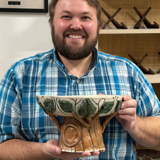 A member holding a pedestal dish with green leaves along the rim and a base carved like a tree trunk at The Clay Hole pottery studio in Draper, Utah
