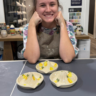 A member resting chin on fists behind three cream dishes with ridged centers and painted yellow lemons at The Clay Hole pottery studio in Draper, Utah