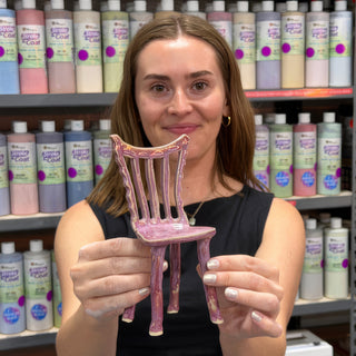 A member holding up a tiny spindle back chair with four legs in glossy pink purple glaze at The Clay Hole pottery studio in Draper, Utah
