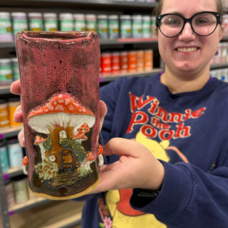 A member holding a tall rectangular vase with speckled red glaze and a raised red mushroom house at The Clay Hole pottery studio in Draper, Utah
