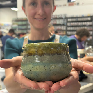 A member holding a small rounded bowl with an olive green top band and a speckled blue gray lower half at The Clay Hole pottery studio in Draper, Utah