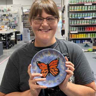 A member holding a round plate with an orange and black butterfly on a periwinkle blue glaze at The Clay Hole pottery studio in Draper, Utah