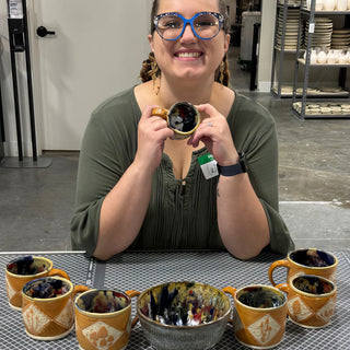 A member holding a small cup behind several orange mugs with carved diamond panels and a gray bowl at The Clay Hole pottery studio in Draper, Utah