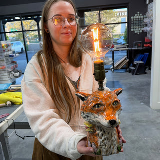 A member holding a lamp base shaped like an orange and white fox head with a lit bulb on top at The Clay Hole pottery studio in Draper, Utah