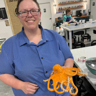 A member holding a bright orange glossy octopus sculpture with curled tentacles and cream colored suckers at The Clay Hole pottery studio in Draper, Utah