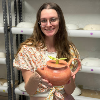 A member holding a salmon orange teapot with a loop handle and a yellow green leaf on the lid at The Clay Hole pottery studio in Draper, Utah
