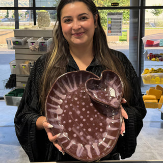 A member holding an oval plum brown platter with white dots, striped edges and a small heart shaped dish at The Clay Hole pottery studio in Draper, Utah