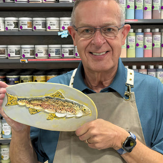 A member holding an oval pale yellow platter with a raised spotted fish in yellow, orange and brown at The Clay Hole pottery studio in Draper, Utah