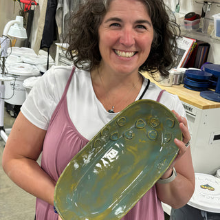 A member holding a long oval platter in glossy teal and olive glaze with small outlined shapes along one edge at The Clay Hole pottery studio in Draper, Utah