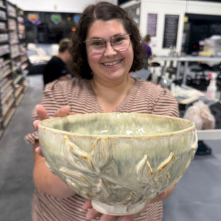 A member holding out a wide pale green bowl with raised leaves and stems on its outer wall at The Clay Hole pottery studio in Draper, Utah